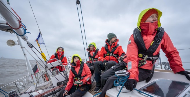 auf einem Segelschiff in Schräglage nach links, die fünf Segler sitzen entspannt am Rand und schaffen Ausgleich