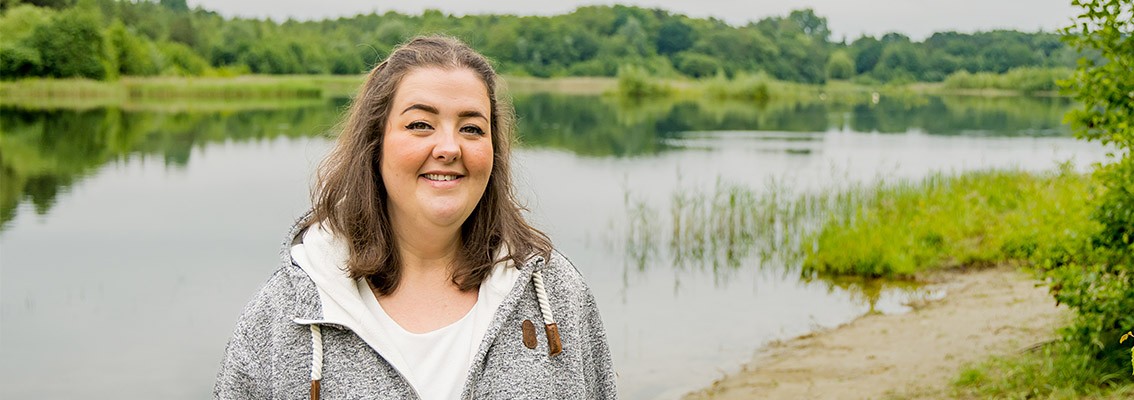 Junge Frau, die an einem See steht, lächelt freundlich in die Kamera 